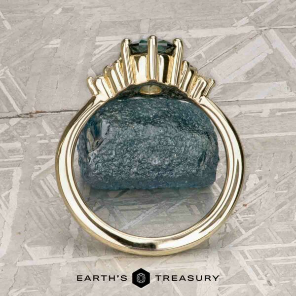 The “Mari” ring in 14k yellow gold with 1.63-Carat Montana Sapphire The “Mari” ring in 14k yellow gold with a 1.63-carat Montana Sapphire is shown on a rough blue stone, with the "Earth's Treasury" logo and text at the bottom.