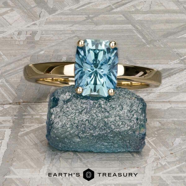 The "Freya" ring in 14k yellow gold with 2.27-Carat Montana Sapphire The "Freya" ring in 14k yellow gold with a 2.27-carat Montana sapphire rests on a rough blue stone atop a textured gray surface. The Earth’s Treasury logo appears at the bottom.
