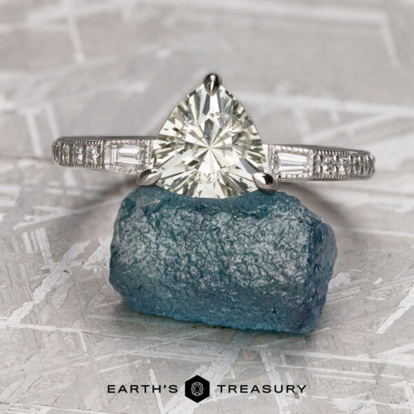 The “Adelaide” ring in 14k white gold with 2.26-Carat Montana Sapphire The “Adelaide” ring in 14k white gold with a 2.26-carat Montana sapphire is displayed on a blue rough stone, set against a geometric background with the "Earth's Treasury" logo below.