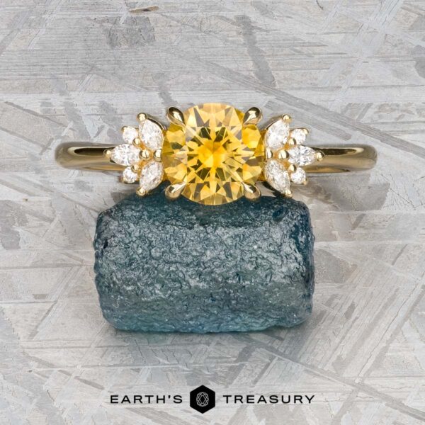 The "Surya" ring in 18k yellow gold with 1.46-carat Montana sapphire The "Surya" ring in 18k yellow gold with a 1.46-carat Montana sapphire is showcased on a rough blue stone, against a textured backdrop with "Earth's Treasury" at the bottom.