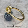 The "Surya" ring in 18k yellow gold with a 1.46-carat Montana sapphire is displayed resting against a rough, dark mineral on a textured surface.