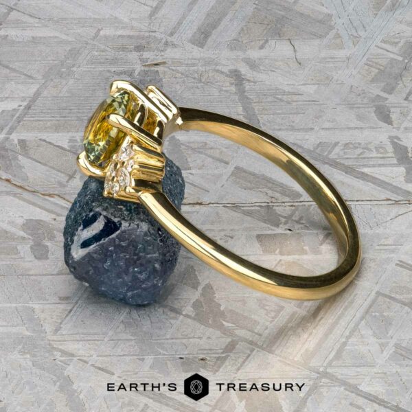 The "Surya" ring in 18k yellow gold with 1.46-carat Montana sapphire The "Surya" ring in 18k yellow gold with a 1.46-carat Montana sapphire is displayed resting against a rough, dark mineral on a textured surface.