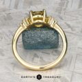 The "Surya" ring in 18k yellow gold with a 1.46-carat Montana sapphire is displayed upright before a rough blue crystal on a textured gray surface. "EARTH'S TREASURY" is shown at the bottom.