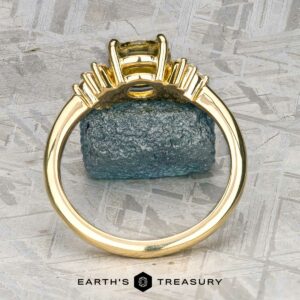 The "Surya" ring in 18k yellow gold with a 1.46-carat Montana sapphire is displayed upright before a rough blue crystal on a textured gray surface. "EARTH'S TREASURY" is shown at the bottom.