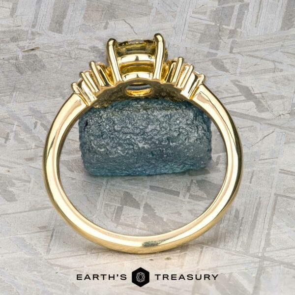 The "Surya" ring in 18k yellow gold with 1.46-carat Montana sapphire The "Surya" ring in 18k yellow gold with a 1.46-carat Montana sapphire is displayed upright before a rough blue crystal on a textured gray surface. "EARTH'S TREASURY" is shown at the bottom.