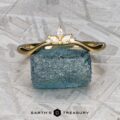 The "Surya" band in 18k yellow gold, featuring a floral motif, is set atop a rough blue gemstone on a textured gray surface with the "Earth's Treasury" logo below.