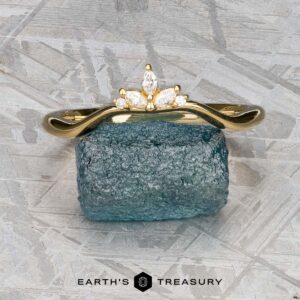 The "Surya" band in 18k yellow gold, featuring a floral motif, is set atop a rough blue gemstone on a textured gray surface with the "Earth's Treasury" logo below.