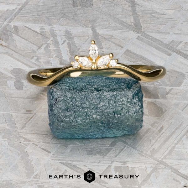 The "Surya" band in 18k yellow gold The "Surya" band in 18k yellow gold, featuring a floral motif, is set atop a rough blue gemstone on a textured gray surface with the "Earth's Treasury" logo below.