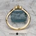 The "Surya" band in 18k yellow gold stands upright before a rough blue gemstone on a textured gray background, with the "Earth’s Treasury" logo displayed at the bottom.