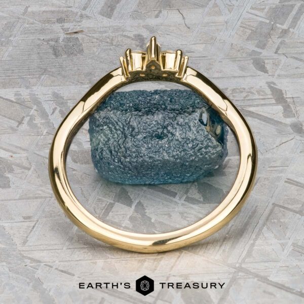The "Surya" band in 18k yellow gold The "Surya" band in 18k yellow gold stands upright before a rough blue gemstone on a textured gray background, with the "Earth’s Treasury" logo displayed at the bottom.