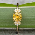 The "Surya" ring in 18k yellow gold features a 1.46-carat Montana sapphire at the center, accented by clusters of small white stones, and is displayed on a green leaf with the "Earth's Treasury" logo below.