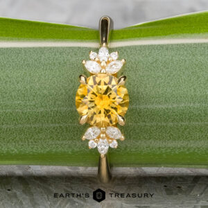 The "Surya" ring in 18k yellow gold features a 1.46-carat Montana sapphire at the center, accented by clusters of small white stones, and is displayed on a green leaf with the "Earth's Treasury" logo below.