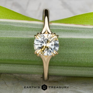 The "Daphne" ring in 14k yellow gold, set with a 1.71-carat Montana sapphire, stands upright against a green leaf background. The Earth's Treasury logo is shown below.
