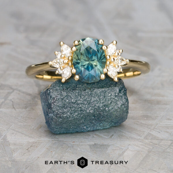 The "Surya" ring in 18k yellow gold with 1.69-carat Montana sapphire The "Surya" ring in 18k yellow gold with a 1.69-carat Montana sapphire rests elegantly on a rough blue-green mineral atop a gray surface.