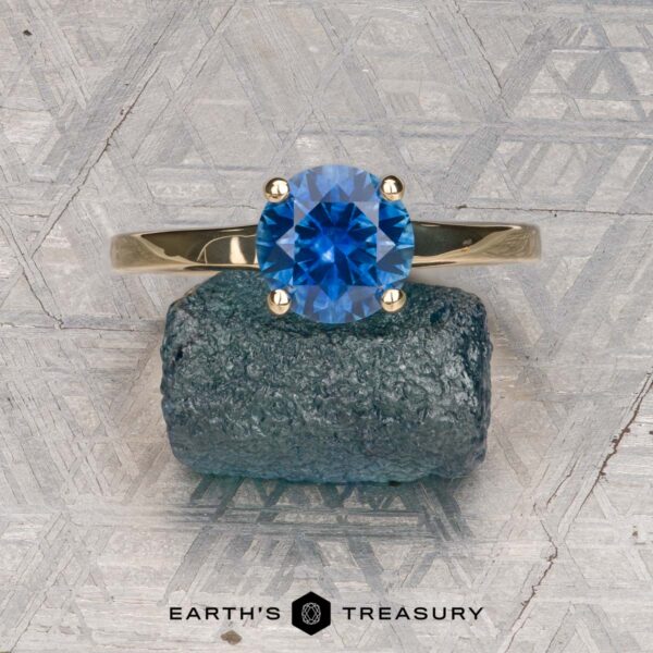 The "Freya" ring in 14k yellow gold with 1.51-carat Montana sapphire The "Freya" ring in 14k yellow gold with a 1.51-carat Montana sapphire is balanced on a rough blue crystal, set against a patterned grey background, with the Earth's Treasury logo at the bottom.