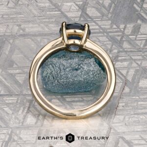 The "Freya" ring in 14k yellow gold with a 1.51-carat Montana sapphire is shown upright on rough blue mineral, with a patterned metallic background and the "Earth's Treasury" logo at the bottom.