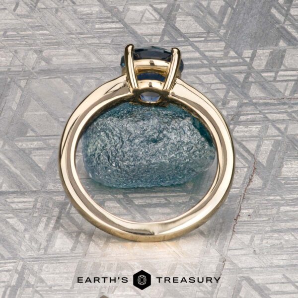 The "Freya" ring in 14k yellow gold with 1.51-carat Montana sapphire The "Freya" ring in 14k yellow gold with a 1.51-carat Montana sapphire is shown upright on rough blue mineral, with a patterned metallic background and the "Earth's Treasury" logo at the bottom.