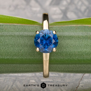 A 14k yellow gold "Freya" ring with a 1.51-carat Montana sapphire, shown on a green leaf background. "Earth's Treasury" logo at the bottom.