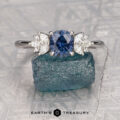 The "Surya" ring in 14k white gold with a 1.08-carat Montana sapphire, shown with side diamonds and resting on a rough blue gemstone. “Earth’s Treasury” logo displayed at the bottom.