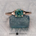 The "Daphne" ring, crafted in 14K rose gold with a 1.06-carat Australian sapphire, rests atop a rough blue stone on a textured surface. "EARTH'S TREASURY" is displayed below—design inspired by the Daphne collection.
