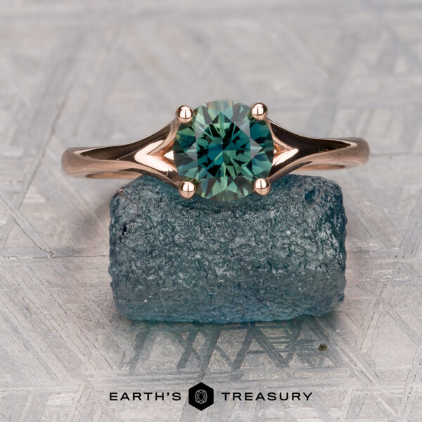 The "Daphne" ring in 14K rose gold with 1.06-carat Australian sapphire The "Daphne" ring, crafted in 14K rose gold with a 1.06-carat Australian sapphire, rests atop a rough blue stone on a textured surface. "EARTH'S TREASURY" is displayed below—design inspired by the Daphne collection.