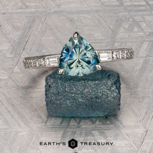 Roe, Ana The Roe, Ana ring—silver with a triangular blue gemstone and side diamonds—is displayed on a rough blue stone with a textured background. "EARTH'S TREASURY" appears at the bottom.