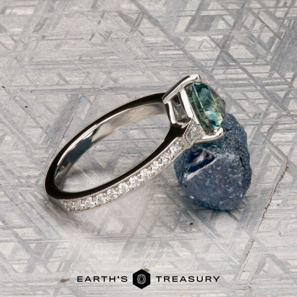 Roe, Ana Roe, Ana ring with a green gemstone and diamond accents rests on a rough blue stone atop a patterned gray surface. Earth’s Treasury logo is visible at the bottom.