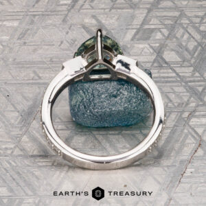 A Roe, Ana Ring featuring a silver band with a green gemstone stands upright on a textured metallic surface, with "EARTH'S TREASURY" displayed below.