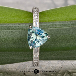 A silver Roe, Ana ring featuring a triangular blue gemstone and small side diamonds rests on green leaves; the "Earth's Treasury" logo appears below.