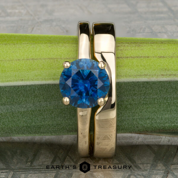 The "Freya" ring in 14K yellow gold with 1.51-carat Montana sapphire, alongside the Custom-Fit Notched Band in 14K yellow gold The "Freya" ring in 14K yellow gold with a 1.51-carat Montana sapphire sits beside the Custom-Fit Notched Band in matching gold, both displayed on a green stem.