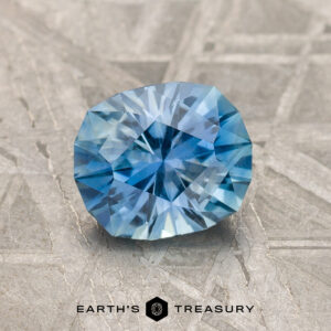 A 1.39-Carat Medium Blue Montana Sapphire with a round, faceted cut is displayed on a textured gray surface featuring the "Earth's Treasury" logo and text at the bottom.