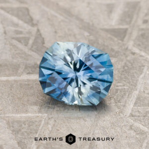 A 1.04-carat medium blue-sky blue particolored Montana sapphire (H) rests on a textured gray surface featuring the "Earth's Treasury" logo and text at the bottom.
