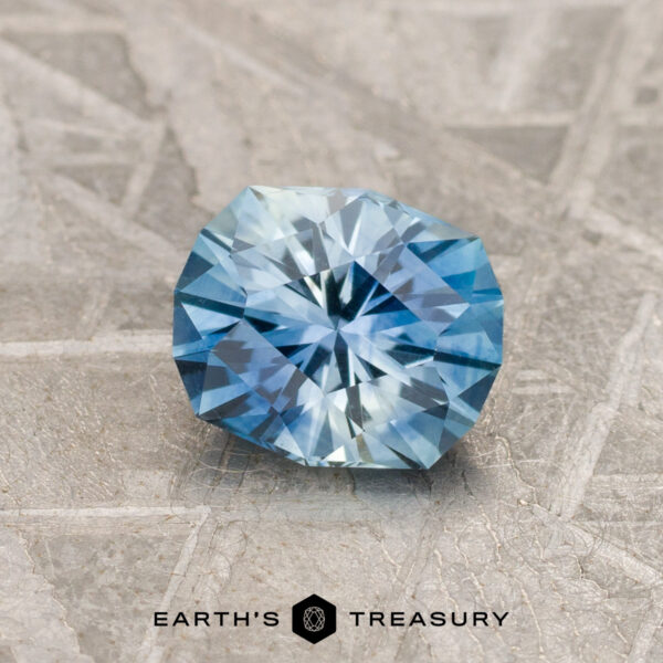 1.04-Carat Medium Blue-Sky Blue Particolored Montana Sapphire (H A 1.04-carat medium blue-sky blue particolored Montana sapphire (H) rests on a textured gray surface featuring the "Earth's Treasury" logo and text at the bottom.