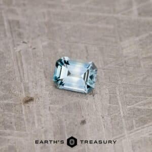 A rectangular, emerald-cut 1.21-carat teal blue Montana sapphire sits on a textured gray surface. The gemstone's facets catch the light, highlighting its clarity and precision. The words "Earth's Treasury" are printed at the bottom.