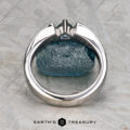 The Stepahin, Sara ring with a central gemstone stands upright before a rough blue stone on a textured gray surface. The "Earth's Treasury" logo is shown at the bottom.