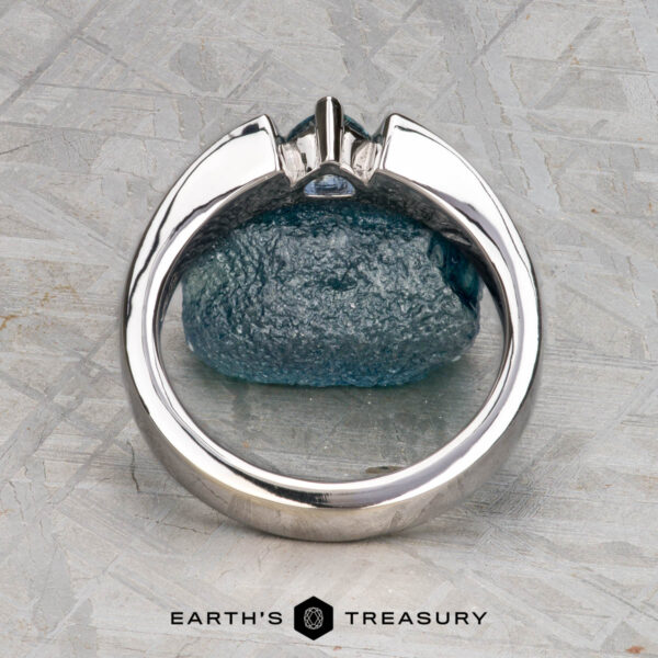 The "Fuji" ring in 14K white gold The Stepahin, Sara ring with a central gemstone stands upright before a rough blue stone on a textured gray surface. The "Earth's Treasury" logo is shown at the bottom.