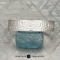 The "Rainier" ring in platinum is displayed upright on a rough blue stone, with a geometric patterned background and the "Earth's Treasury" logo below.