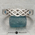 The "Rowan" ring in 14K white gold features a Celtic knot design with blue gemstones on each side, shown on a rough blue stone with the Earth's Treasury logo below.