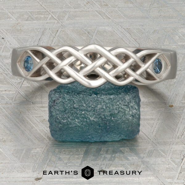 The "Rowan" ring in 14K white gold The "Rowan" ring in 14K white gold features a Celtic knot design with blue gemstones on each side, shown on a rough blue stone with the Earth's Treasury logo below.