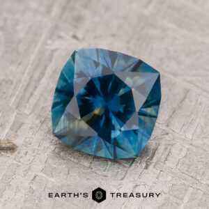 A 1.62-carat rich blue-green-yellow particolored Montana Sapphire (heated) with a square cushion cut rests on a textured gray surface. The "EARTH'S TREASURY" logo is visible at the bottom.