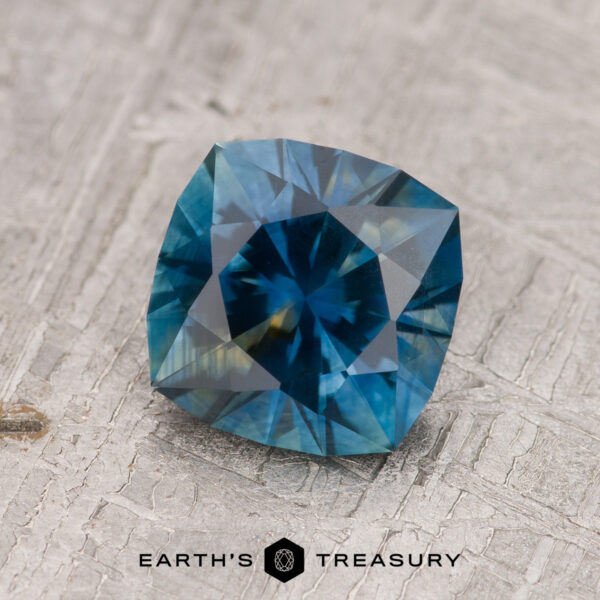 1.62-Carat Rich Blue-Green-Yellow Particolored Montana Sapphire (Heated) A 1.62-carat rich blue-green-yellow particolored Montana Sapphire (heated) with a square cushion cut rests on a textured gray surface. The "EARTH'S TREASURY" logo is visible at the bottom.
