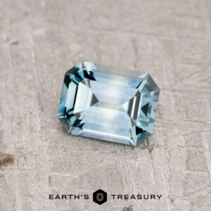 A rectangular, emerald-cut 1.21-carat teal blue Montana sapphire sits on a textured gray surface. The gemstone's facets catch the light, highlighting its clarity and precision. The words "Earth's Treasury" are printed at the bottom.