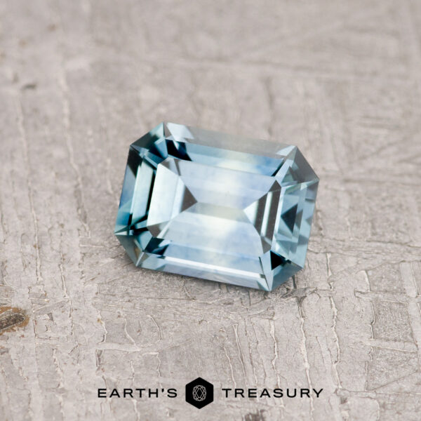 0.64-Carat Aqua Blue Montana Sapphire (Heated) A rectangular, emerald-cut 1.21-carat teal blue Montana sapphire sits on a textured gray surface. The gemstone's facets catch the light, highlighting its clarity and precision. The words "Earth's Treasury" are printed at the bottom.