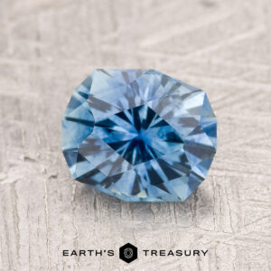 A 1.86-carat teal blue Montana Sapphire (heated) is displayed on a textured light gray surface, with the "Earth's Treasury" logo and text at the bottom.