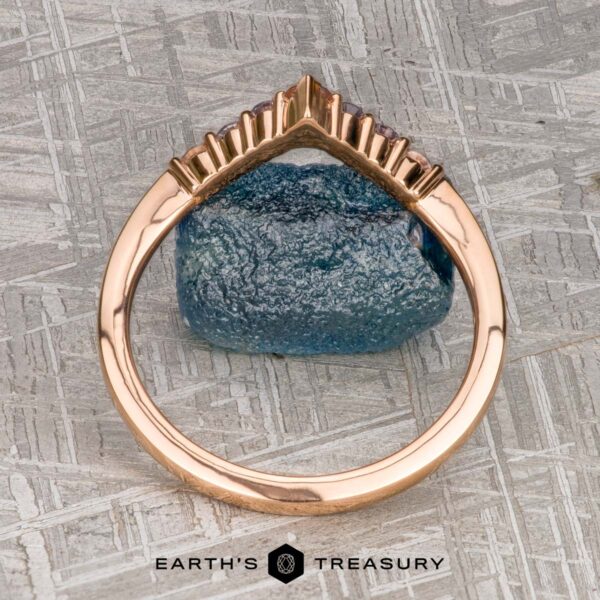 The "Melilai" band in 20k pink gold The "Melilai" band in 20k pink gold, featuring a crown-like design, stands upright against a rough blue gemstone with the Earth's Treasury logo below.