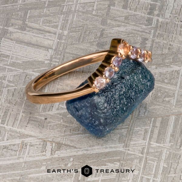The "Melilai" band in 20k pink gold The "Melilai" band in 20k pink gold features a rough blue gemstone and a curved row of small round white stones, displayed on a textured gray surface.