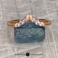 The "Melilai" band in 20k pink gold features seven light pink and lavender gemstones in a chevron design, displayed on a rough blue stone with "EARTH'S TREASURY" below—an elegant piece inspired by harmony.