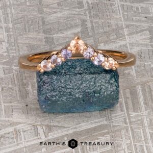 The "Melilai" band in 20k pink gold features seven light pink and lavender gemstones in a chevron design, displayed on a rough blue stone with "EARTH'S TREASURY" below—an elegant piece inspired by harmony.
