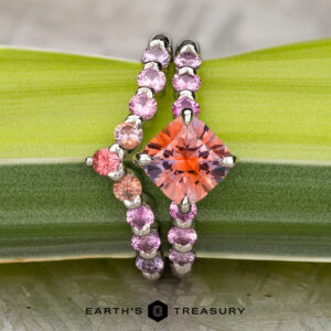 The "Dahlia" ring in platinum with a 2.14-carat orange Montana Sapphire sits beside the matching platinum "Dahlia" band, both shown on a green leaf. The "Earth's Treasury" logo appears at the bottom.