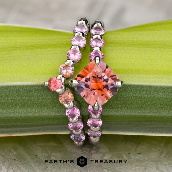 The "Dahlia" ring in platinum with 2.14-Carat Montana Sapphire, alongside the "Dahlia" band in platinum The "Dahlia" ring in platinum with a 2.14-carat orange Montana Sapphire sits beside the matching platinum "Dahlia" band, both shown on a green leaf. The "Earth's Treasury" logo appears at the bottom.
